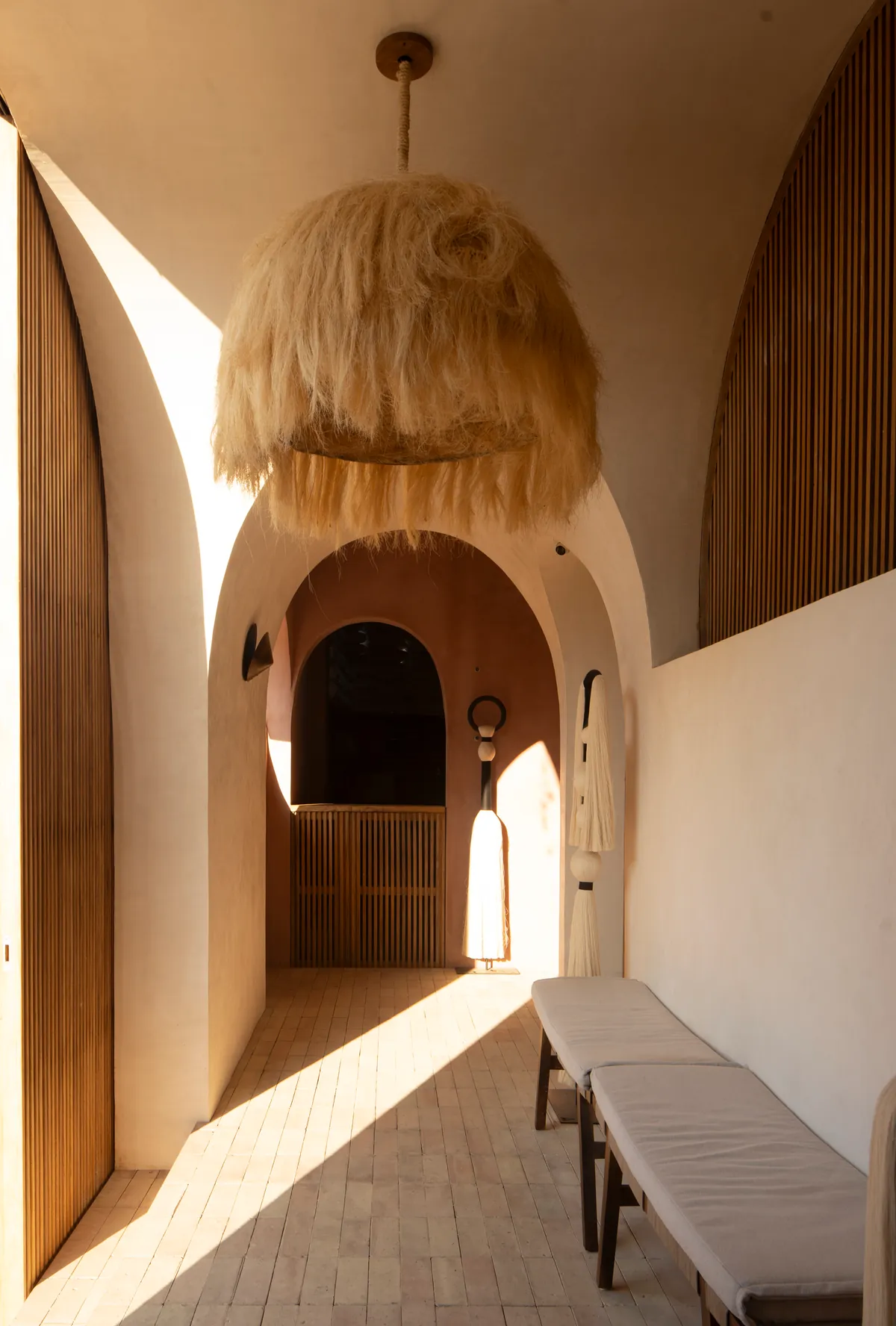 Entrance gallery with nested arches, tall wooden slat doors, a large natural-fibre pendant lamp and benches on a brick floor crossed by a band of sun