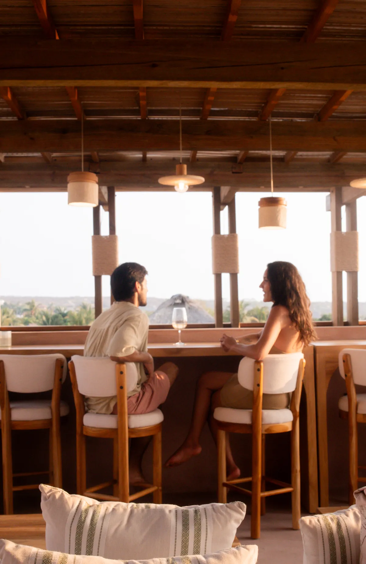 A couple talks at the rooftop’s wooden counter, facing open bays with palm treetops and a thatched palapa roof