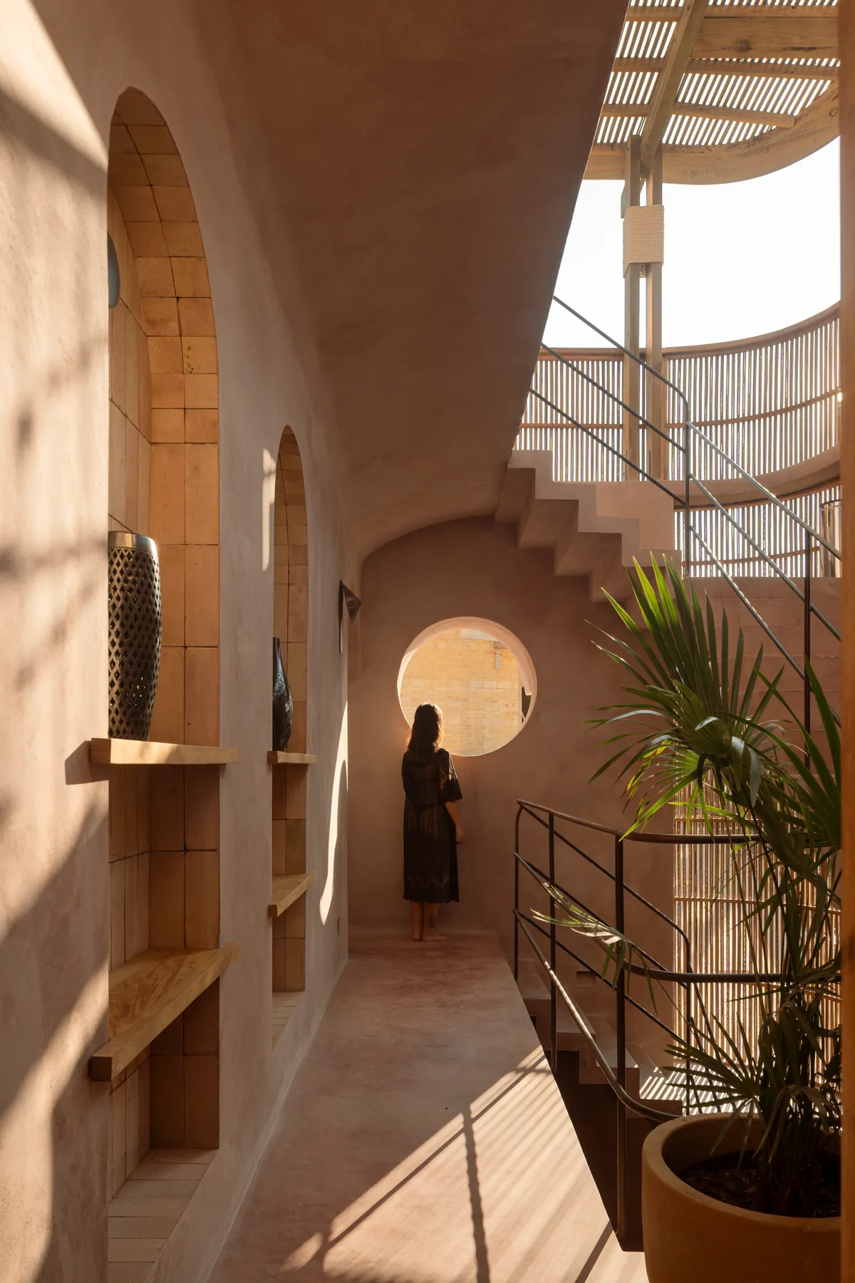 A woman seen from behind at a round window at the end of a pink chukum corridor with brick niches and wooden shelves